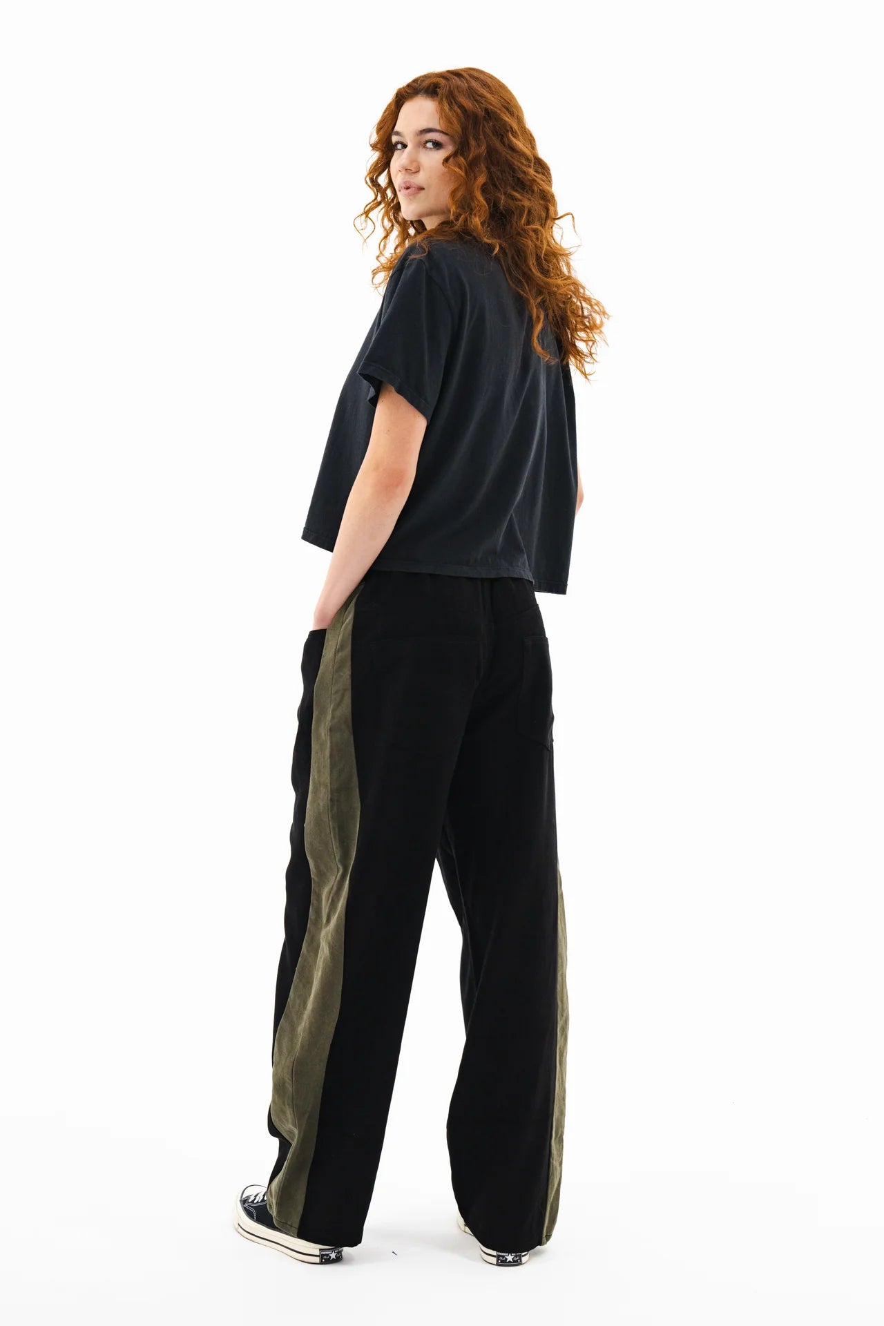 A person with long curly hair wears a loose black T-shirt, ASKK Cancun Pant in black with olive side stripes, and black sneakers, standing and looking over their shoulder against a white background.