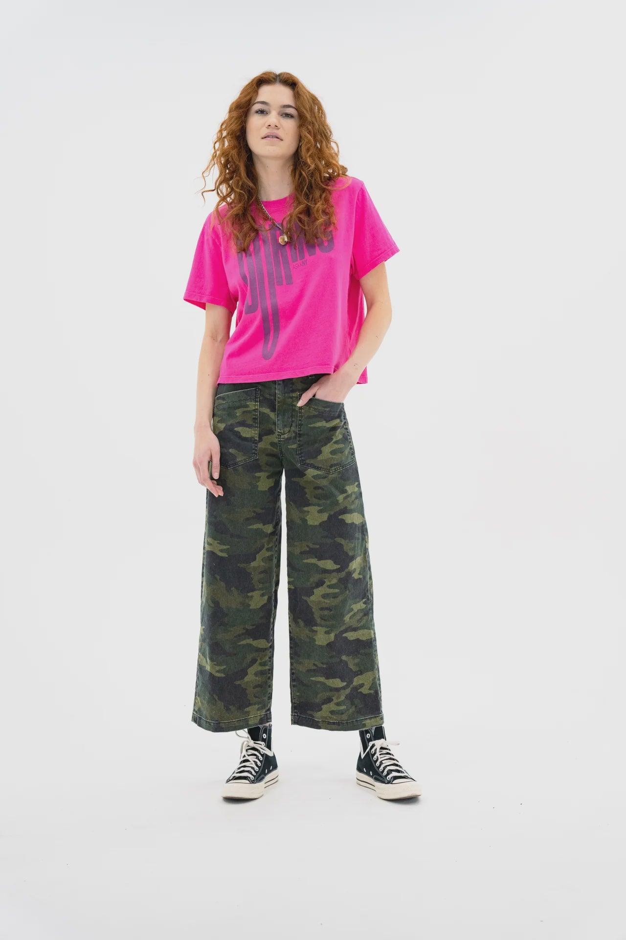 A woman with long curly red hair wears a bright pink graphic tee, ASKK’s Virginia Pant in stretch twill, and black high-top sneakers. She stands confidently against a plain white background.
