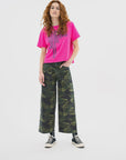 A woman with long curly red hair wears a bright pink graphic tee, ASKK’s Virginia Pant in stretch twill, and black high-top sneakers. She stands confidently against a plain white background.