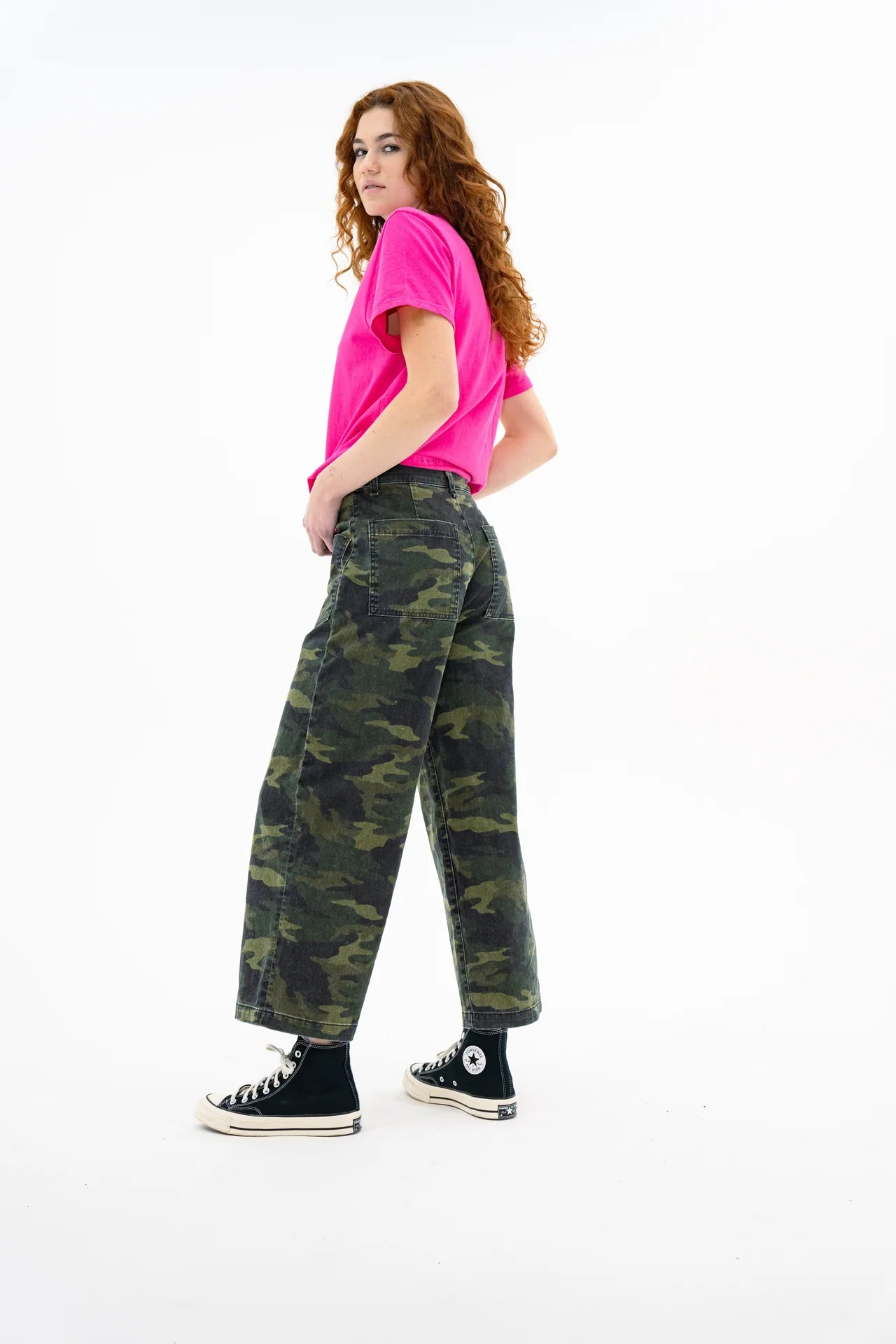 A person with long curly red hair, in a bright pink t-shirt, ASKK's Virginia Pant (cropped wide-leg camo), black socks, and black Converse sneakers stands sideways against a white background.