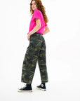 A person with long curly red hair, in a bright pink t-shirt, ASKK's Virginia Pant (cropped wide-leg camo), black socks, and black Converse sneakers stands sideways against a white background.