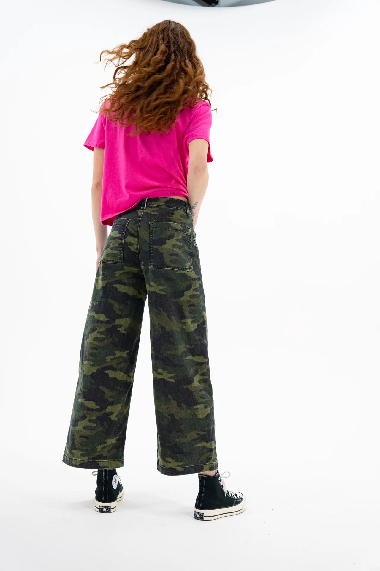 A person with long, wavy hair faces away, wearing a bright pink T-shirt and the ASKK Virginia Pant in camo stretch twill, paired with black high-top sneakers against a white background.