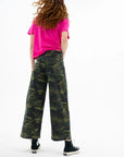 A person with long, wavy hair faces away, wearing a bright pink T-shirt and the ASKK Virginia Pant in camo stretch twill, paired with black high-top sneakers against a white background.
