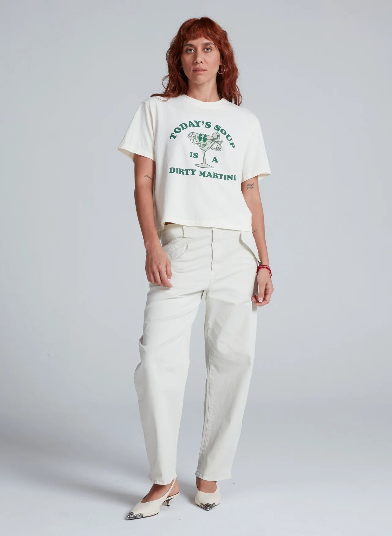 A red-haired person wears the ASKK Shrunken Boy Tee in white with “TODAY&#39;S SOUP IS A DIRTY MARTINI” text, cream pants, and pointed-toe shoes, standing in front of a plain gray background.