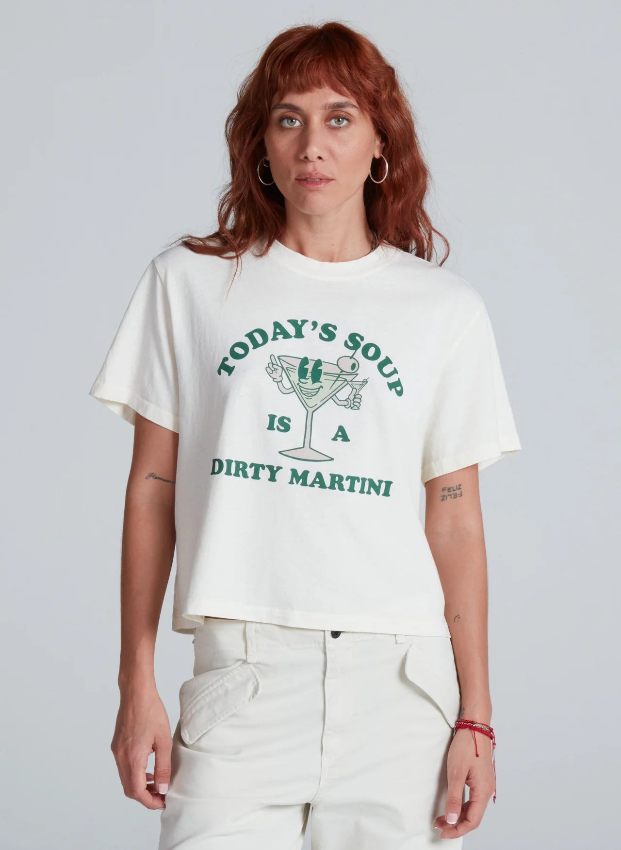 A woman with wavy red hair wears the ASKK Shrunken Boy Tee, a 100% cotton crewneck featuring &quot;TODAY&#39;S SOUP IS A DIRTY MARTINI&quot; and a martini glass graphic. She pairs it with light pants, standing against a plain background.