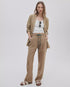A woman poses with her hands in her tan Rag & Bone atlas linen pant, paired with a matching blazer and white tank. She accessorizes with sunglasses hanging from her shirt and completes the look with white sandals against a light backdrop.