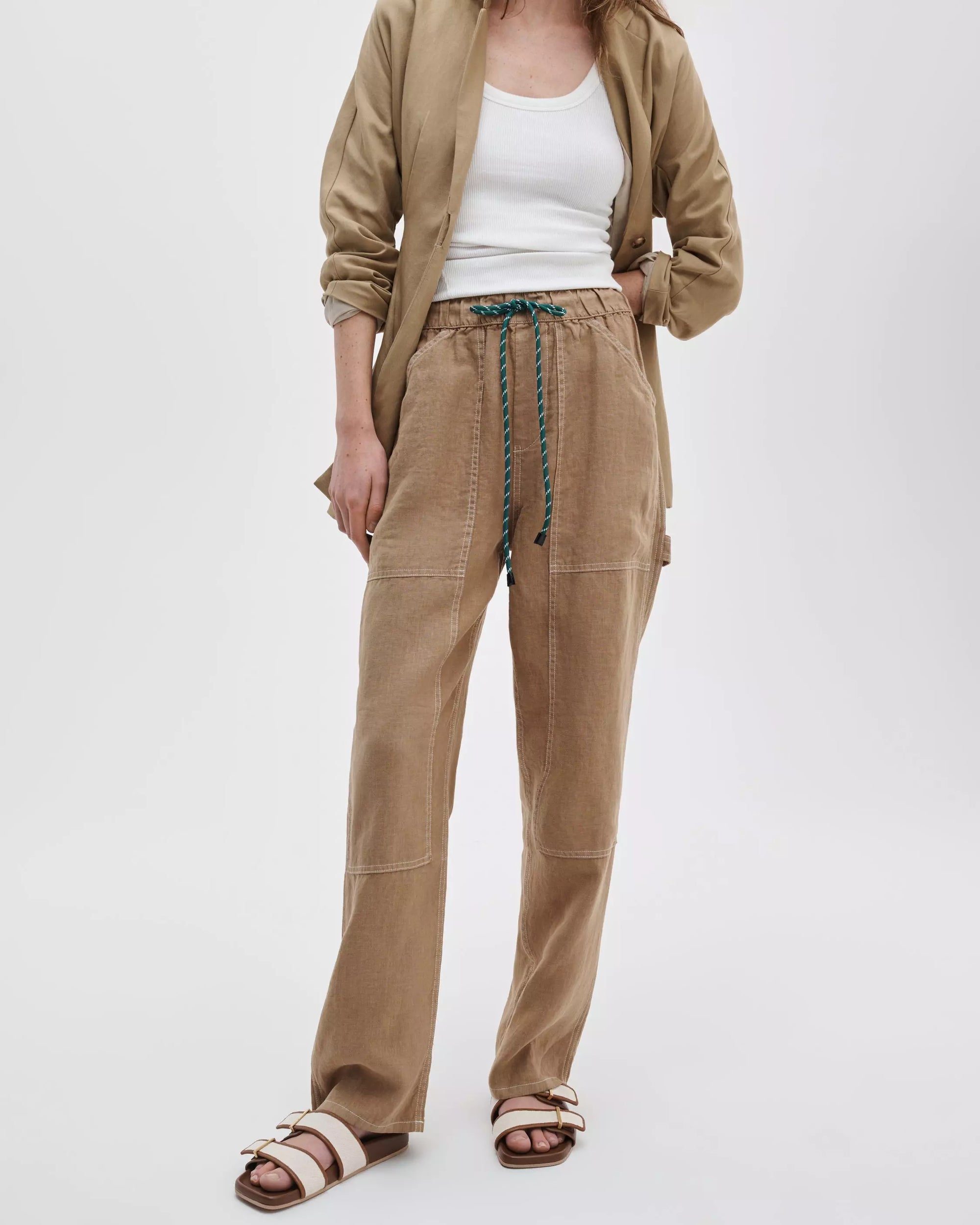 A person stands against a white background wearing a tan jacket over a white tank, Rag & Bone atlas linen pant in a relaxed fit, and beige buckled sandals; their face is not visible.