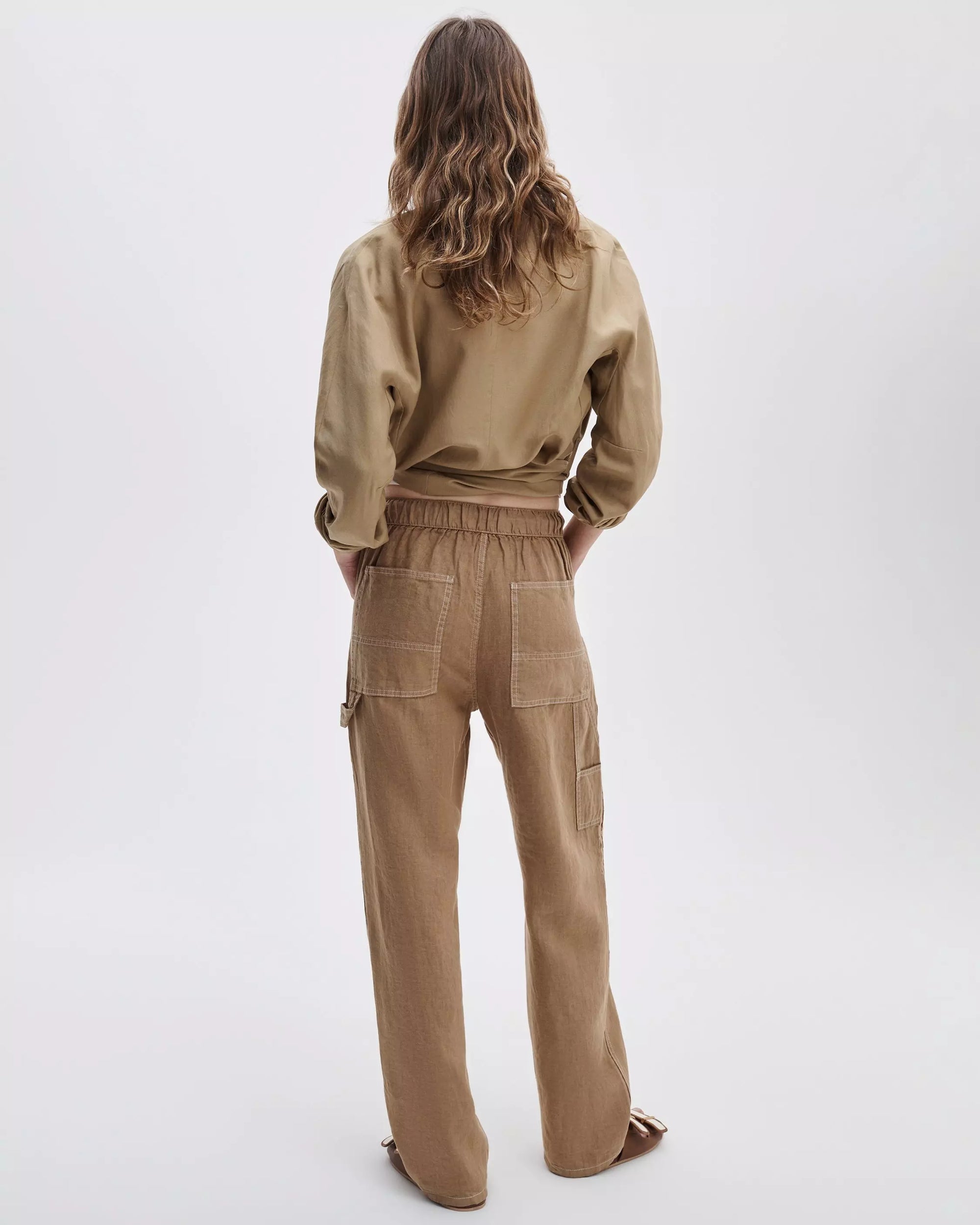 A person with wavy brown hair faces away, wearing a loose tan button-up shirt and relaxed fit high-waisted Rag & Bone atlas linen pant with visible back pockets, set against a plain light background.