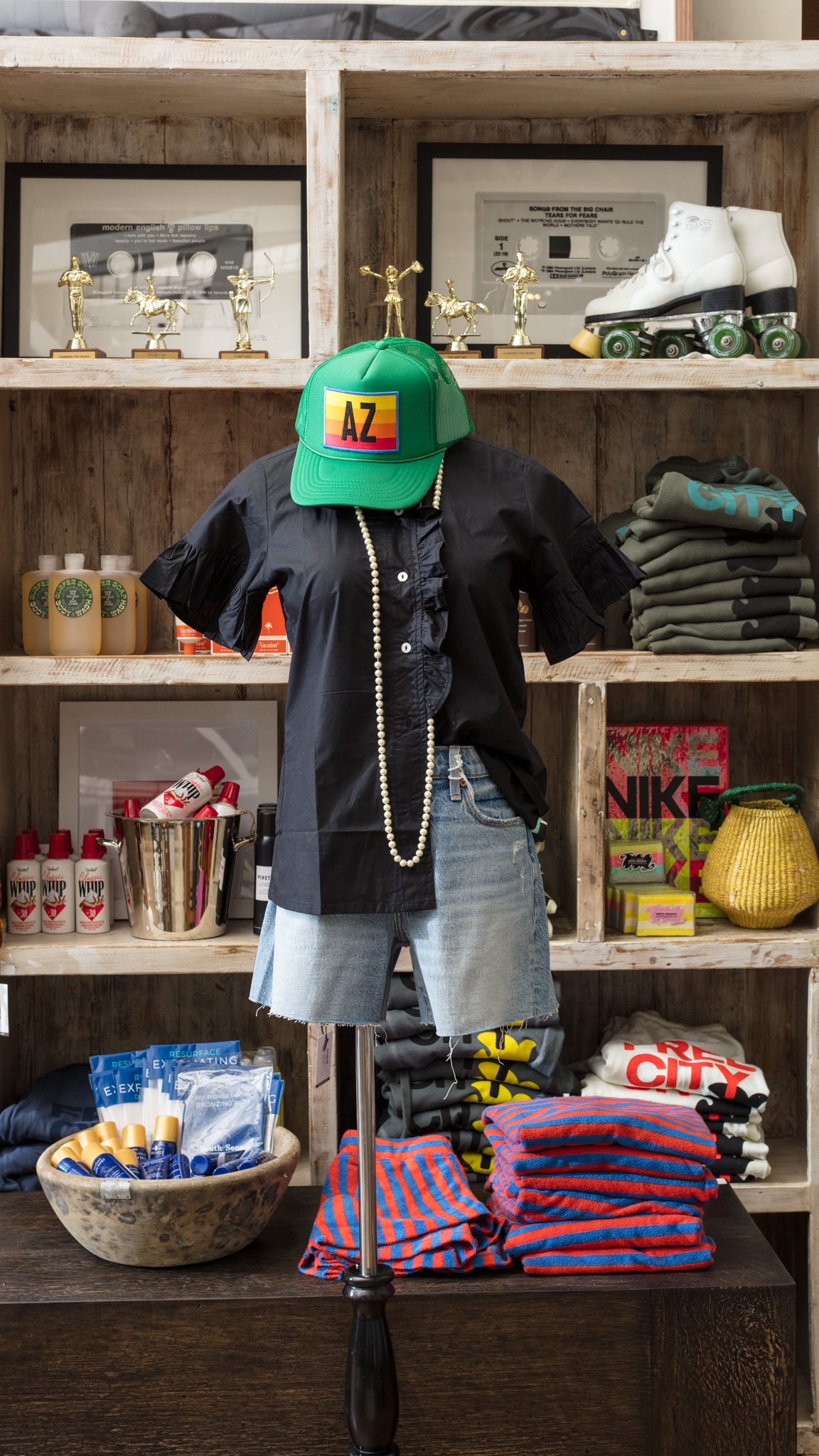 A mannequin displays a black shirt, denim shorts, and an AZ Striped Trucker Hat by Truckers By Tams, exuding Arizona style. A pearl necklace graces its neck. Behind, shelves brim with clothes, roller skates, trophies, Starbucks products, and sports items. Below are folded clothes and grooming products arranged neatly.