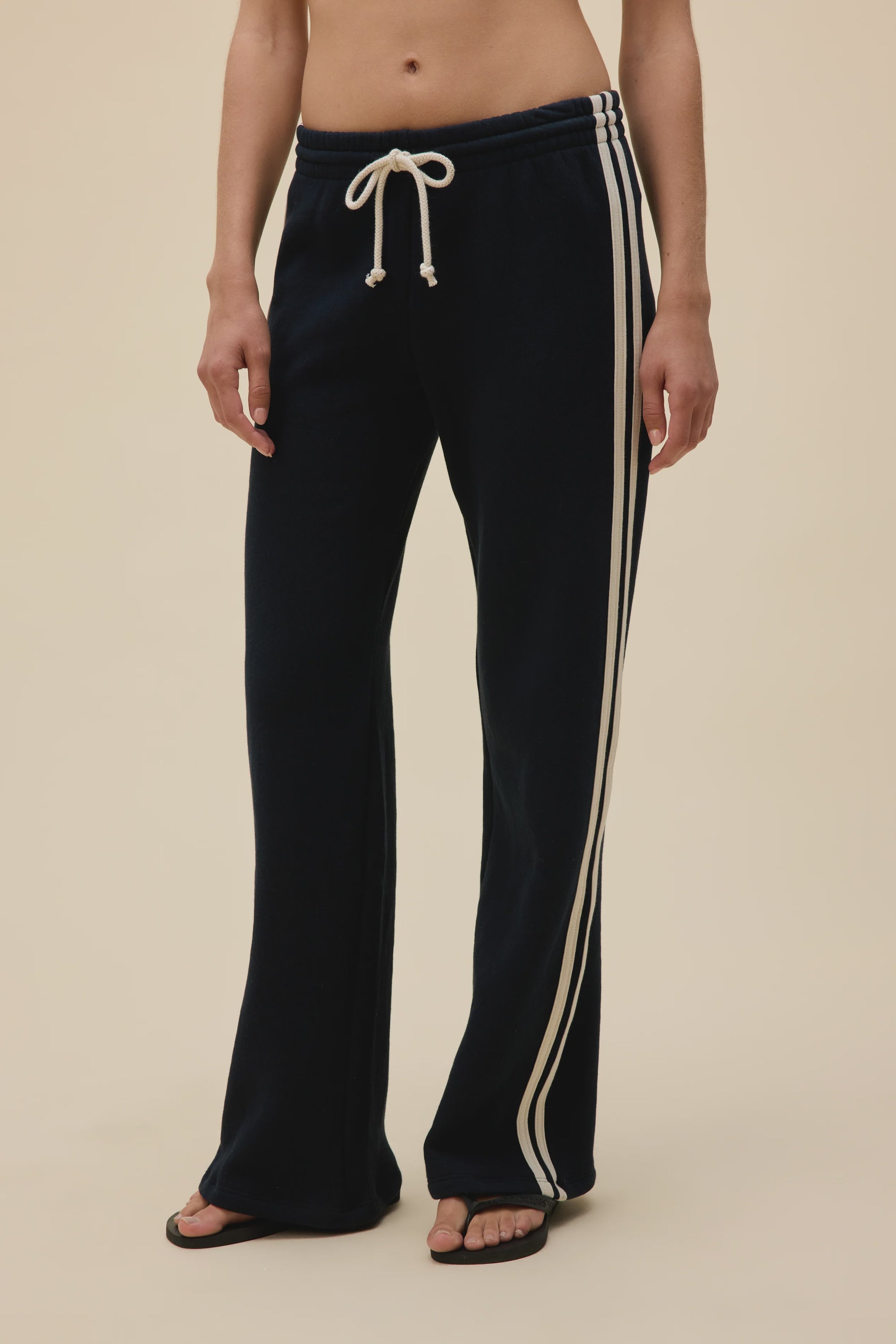 A person wears DayDreamer Track Pant—relaxed fit, black with white side stripes and a drawstring waist—paired with black flip-flops. Their midriff and arms are visible as they stand against a neutral background.