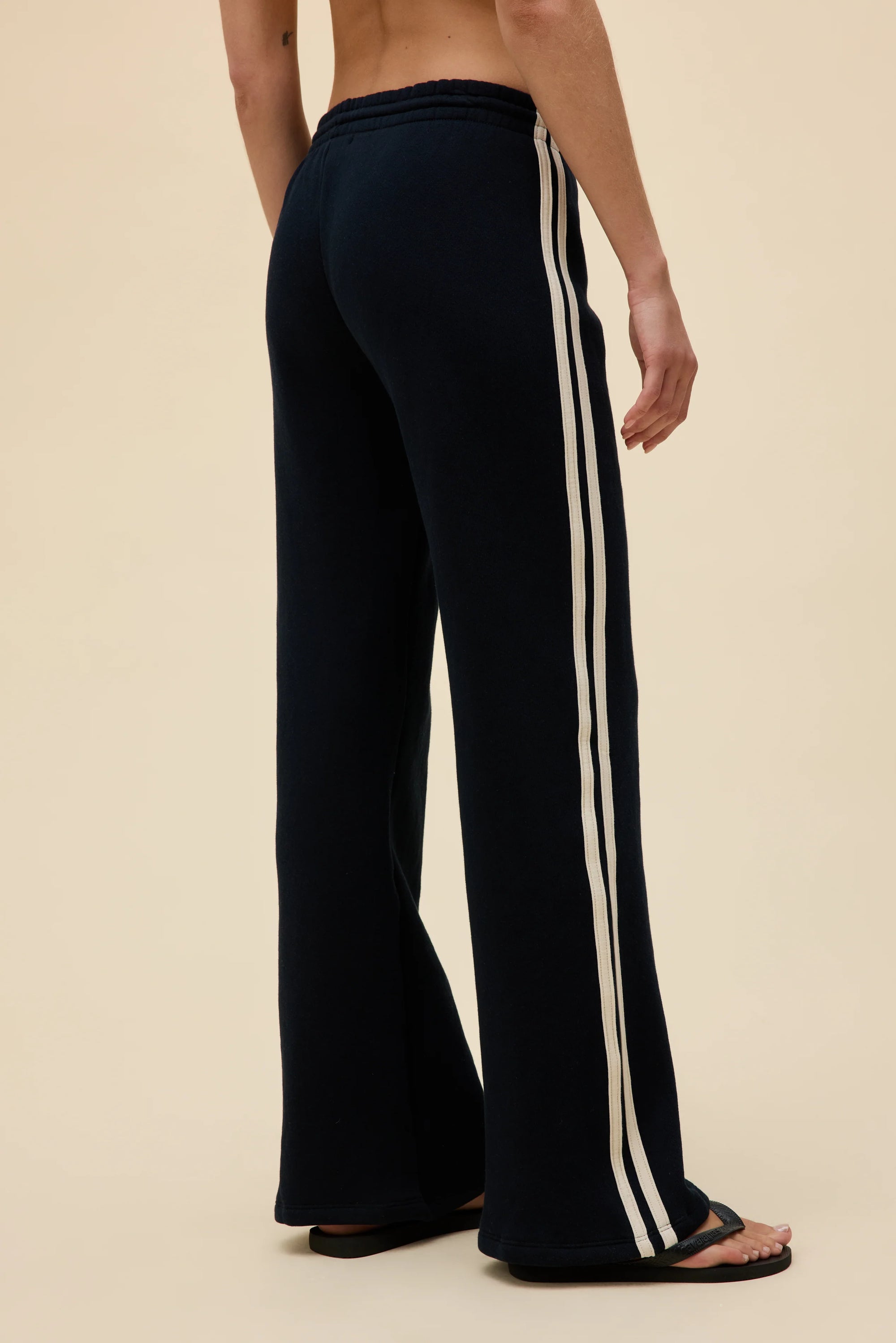 A person models the DayDreamer Track Pant—black relaxed fit with white side stripes—standing barefoot against a beige background, viewed from the back.
