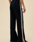 A person models the DayDreamer Track Pant—black relaxed fit with white side stripes—standing barefoot against a beige background, viewed from the back.