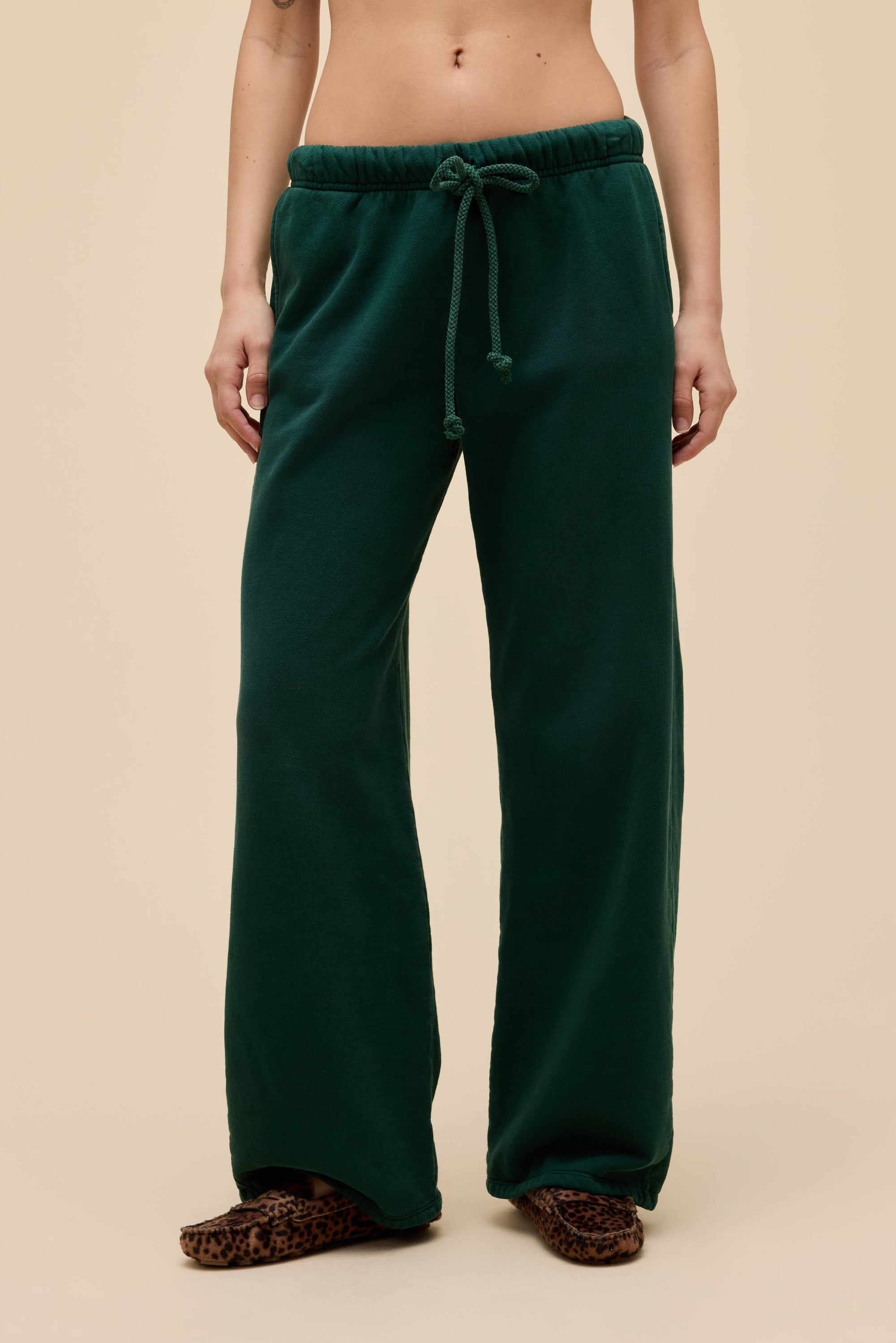 A person wears DayDreamer Drawstring Pants in dark green with a sunfaded effect and side pockets, styled with leopard print slippers, standing against a plain beige background.