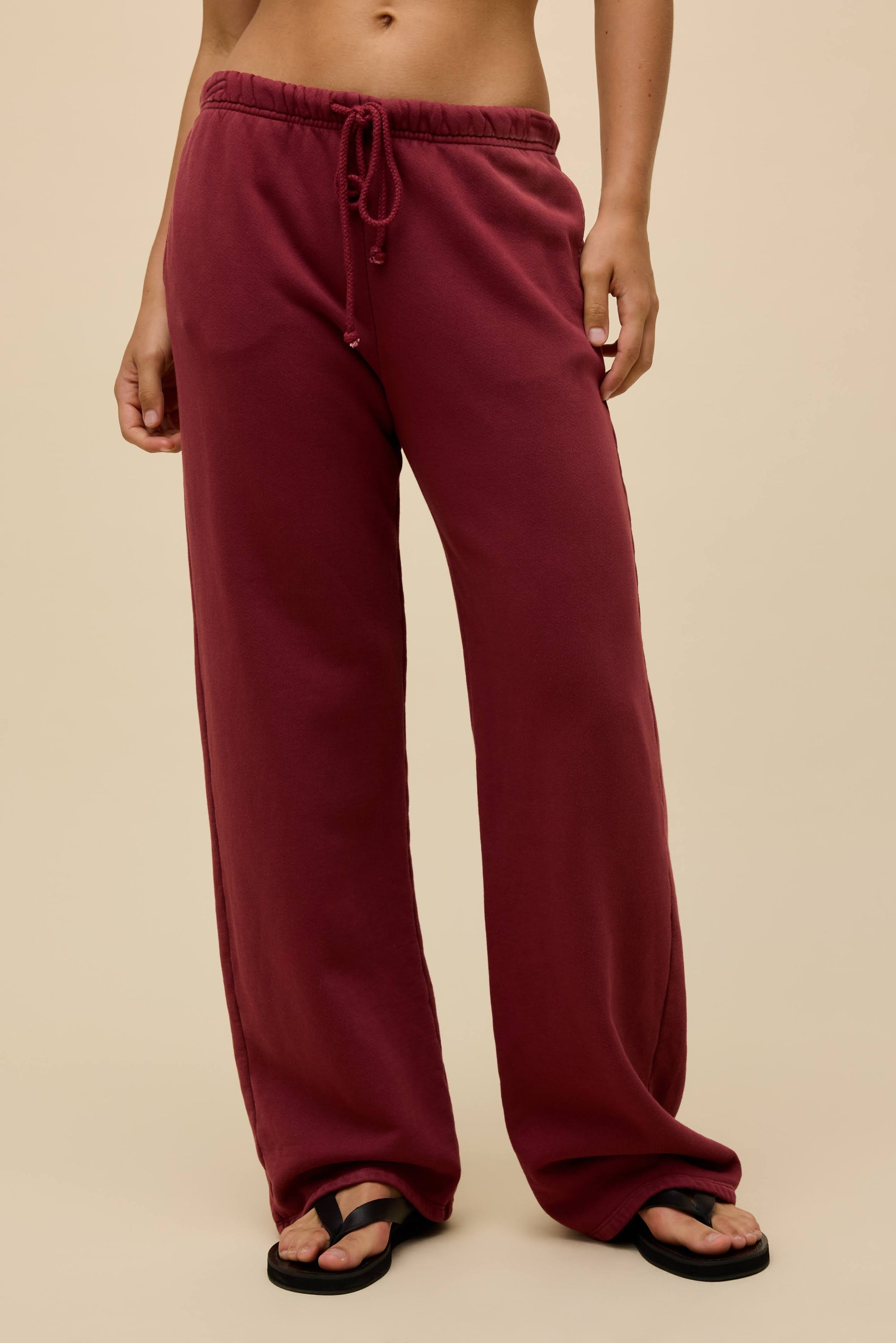 Someone is wearing loose-fitting, high-waisted maroon DayDreamer Drawstring Pants with a sun-faded effect and black flip-flops, standing against a beige background.