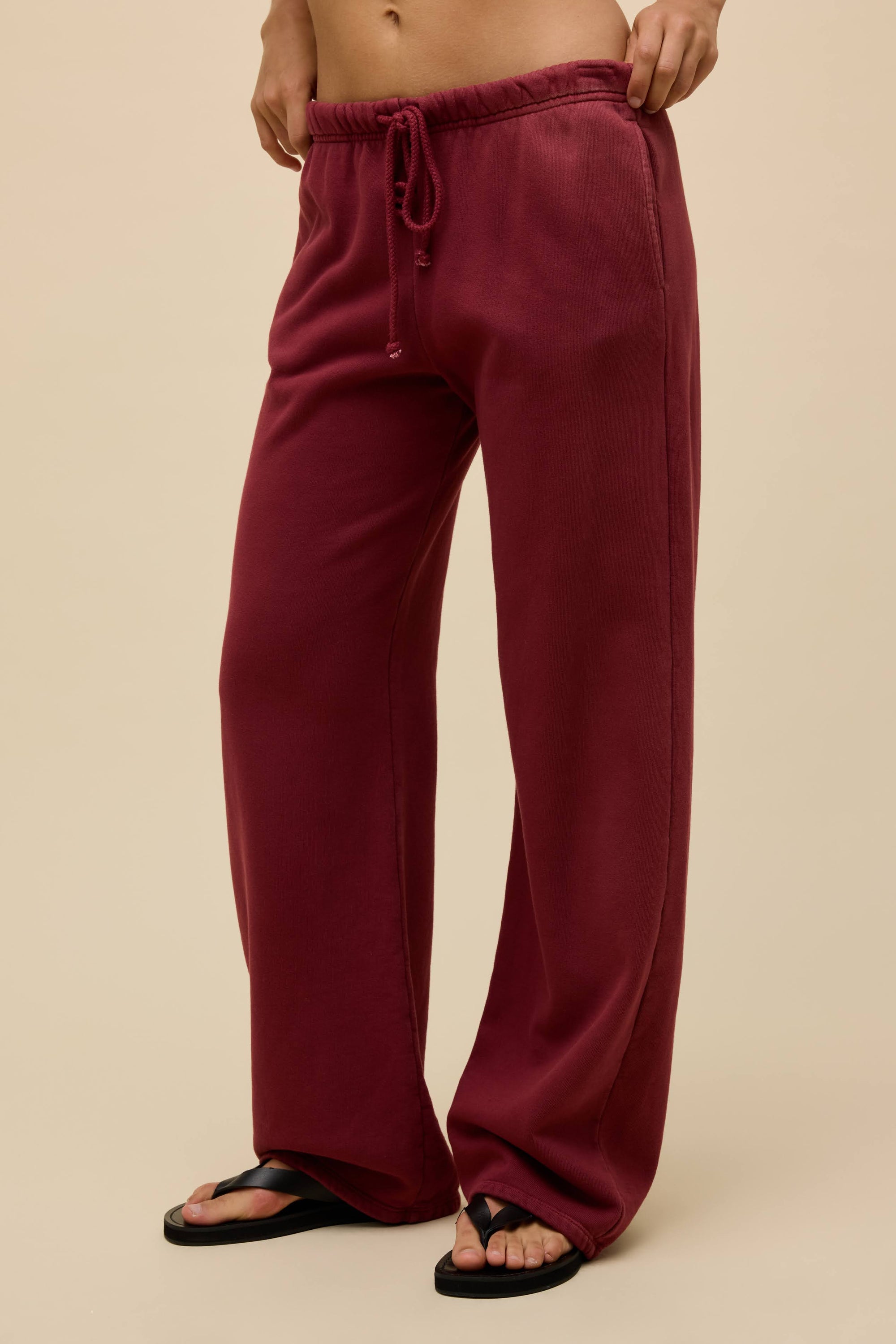 A person models DayDreamer Drawstring Pants—relaxed-fit, straight leg with a sunfaded finish—against a beige background. Only the lower torso and legs are visible, paired with black flip-flops.