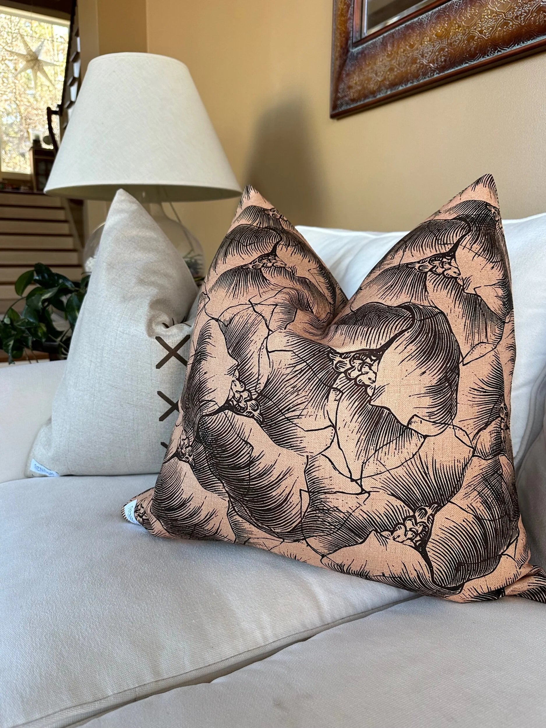 A beige couch is styled with two throw pillows—one solid light color with side lacing, and the other featuring a peach Rennah Collective Milo Pillow 22x22 with a black floral line design. A lamp, framed art, and stairs finish the background.