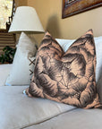 A beige couch is styled with two throw pillows—one solid light color with side lacing, and the other featuring a peach Rennah Collective Milo Pillow 22x22 with a black floral line design. A lamp, framed art, and stairs finish the background.