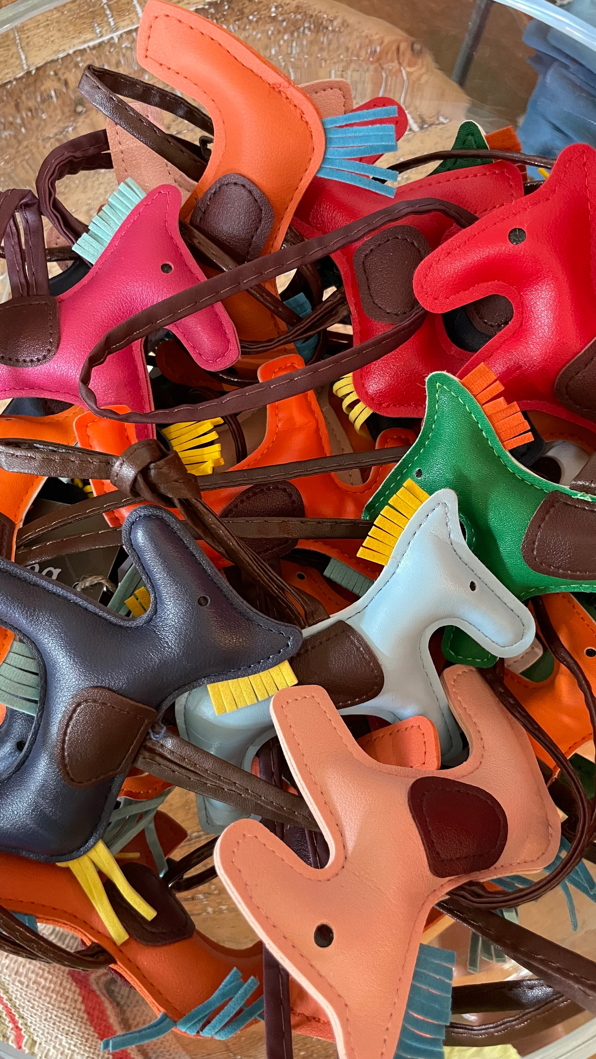 A basket filled with Jeff's Antiques Mini Rodeo Purse Charms, featuring colorful miniature leather horse figures. These charming horses come in various colors including red, green, orange, black, and brown, with some having contrasting saddle and mane colors. The basket is placed on a light wooden surface.