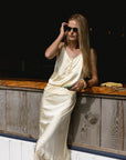 A woman with long blonde hair stands by a wooden counter in bright sunlight, wearing THE SHIRT's The Cami Shirt in cream satin with lace trim, holding sunglasses and a phone—enjoying no blouse gape thanks to the No Gape buttons.