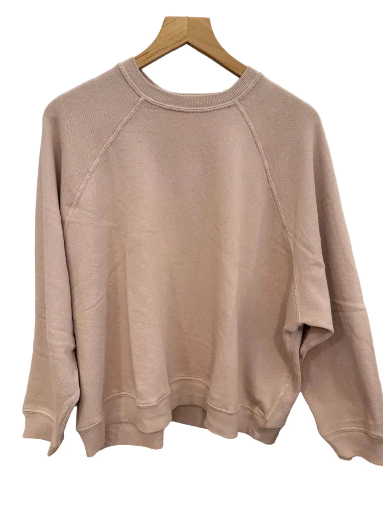 The Rehearsal Sweatshirt by The Great Inc.—a light pink, long-sleeve raglan sweatshirt—hangs on a wooden hanger against a white background, making it the perfect addition to your weekend essentials.