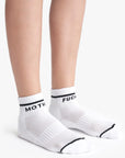 A person wears Mother’s Baby Steps Ankle socks—classic black and white tube socks with bold black stripes and the words “MOTH” and “FUCK” printed in black around the cuffs.