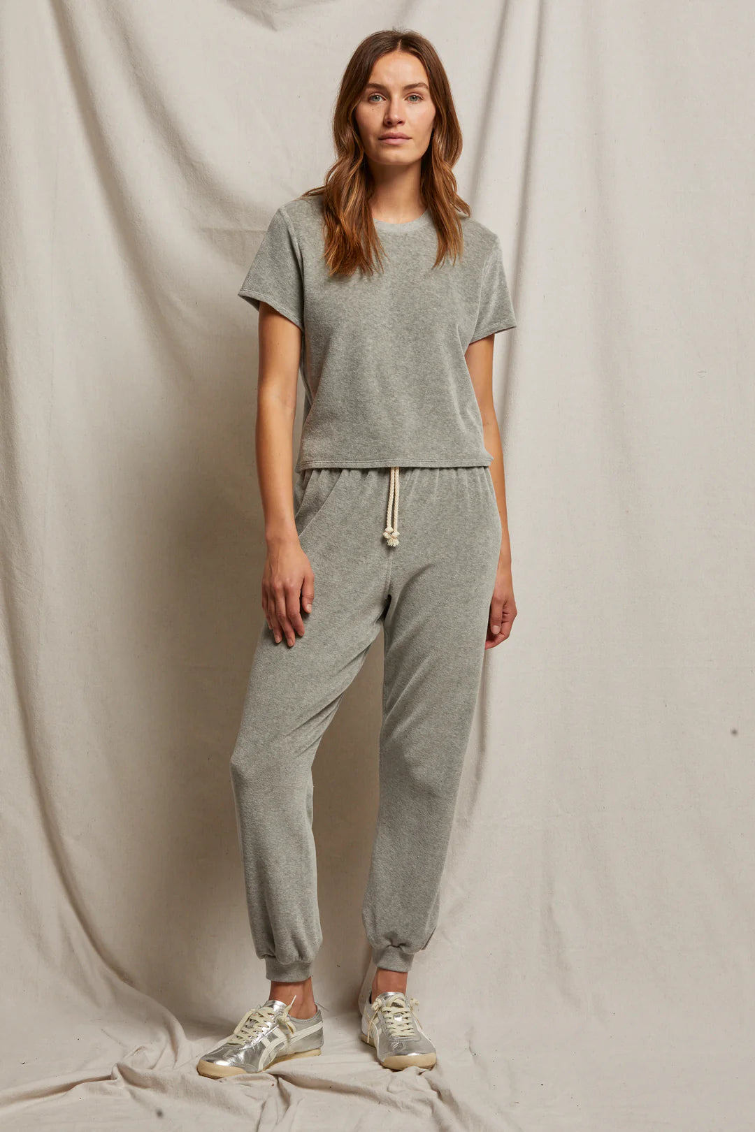 A woman is sporting the Bailey Velour Jogger by Perfectwhitetee in heather grey, paired with sneakers, as she stands in front of a beige fabric backdrop. Her long hair falls naturally as she maintains a relaxed posture.