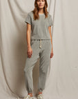 A woman is sporting the Bailey Velour Jogger by Perfectwhitetee in heather grey, paired with sneakers, as she stands in front of a beige fabric backdrop. Her long hair falls naturally as she maintains a relaxed posture.