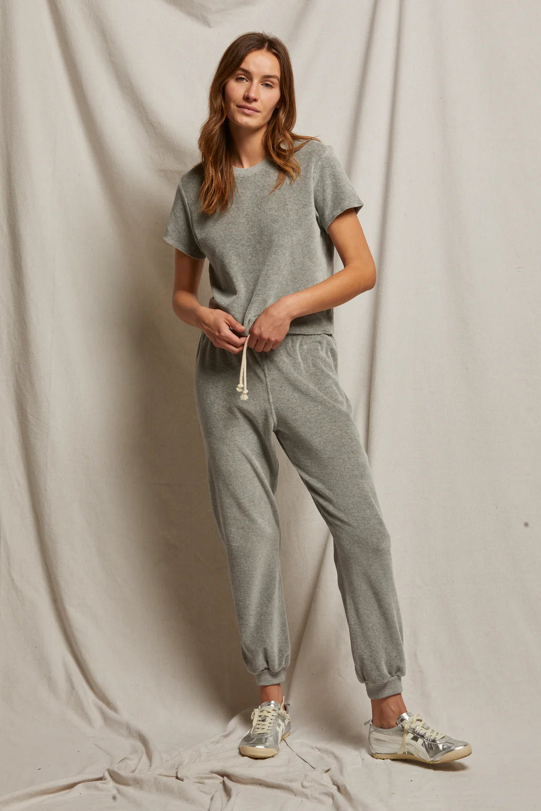 A person poses against a neutral fabric backdrop in a casual heather grey matching set from Perfectwhitetee, featuring the Bailey Velour Jogger and a short-sleeve top. They have long, wavy hair and wear sneakers, creating a comfortable and relaxed look.