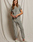 A person poses against a neutral fabric backdrop in a casual heather grey matching set from Perfectwhitetee, featuring the Bailey Velour Jogger and a short-sleeve top. They have long, wavy hair and wear sneakers, creating a comfortable and relaxed look.