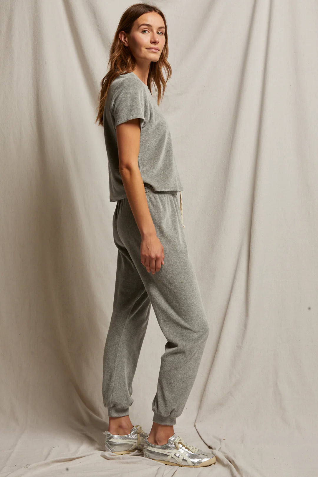 A person stands in profile wearing the Bailey Velour Jogger from Perfectwhitetee along with sneakers, exuding comfort and style. The jogger set complements the simple beige curtain in the background, creating a cohesive and relaxed vibe.