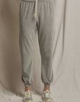 A person is standing in front of a beige fabric backdrop wearing the Bailey Velour Jogger from Perfectwhitetee. These gray joggers feature a drawstring, a relaxed fit, and elastic cuffs. The outfit is completed with matching gray and beige sneakers.