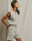 A woman stands before a light fabric backdrop, wearing Perfectwhitetee’s Bari Short and a sleeveless gray top in soft French terry, looking to the side with one hand on her hip.