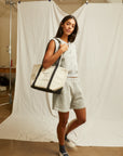A woman stands indoors before a white backdrop, wearing Perfectwhitetee's Bari Short in gray with a matching top, white shoes, dark socks, and carrying a large beige-and-black tote bag on her shoulder.
