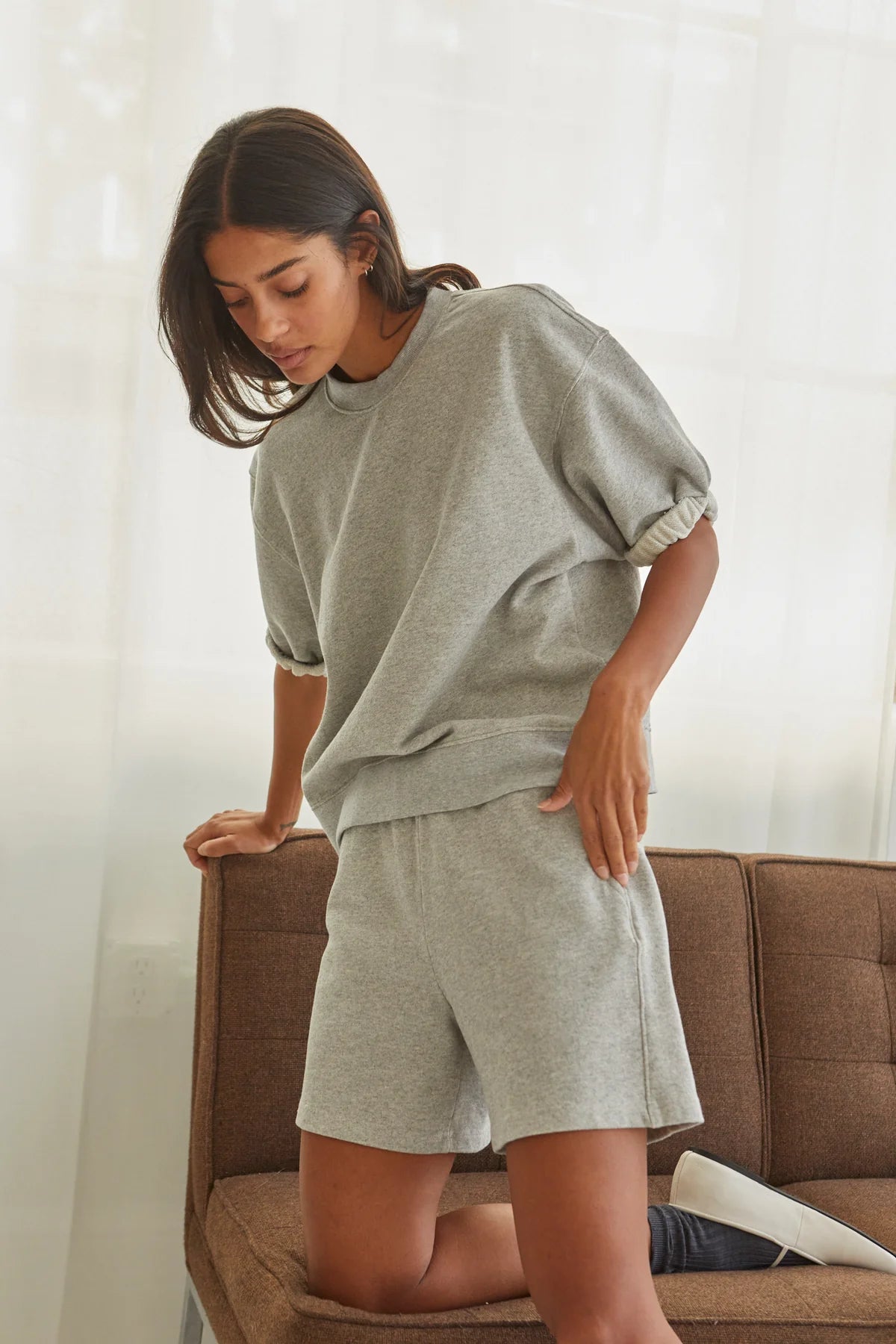 A woman in Perfectwhitetee's Bari Short and a loose gray French terry T-shirt leans on a brown sofa in a bright room with sheer white curtains.