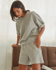 A woman in Perfectwhitetee's Bari Short and a loose gray French terry T-shirt leans on a brown sofa in a bright room with sheer white curtains.