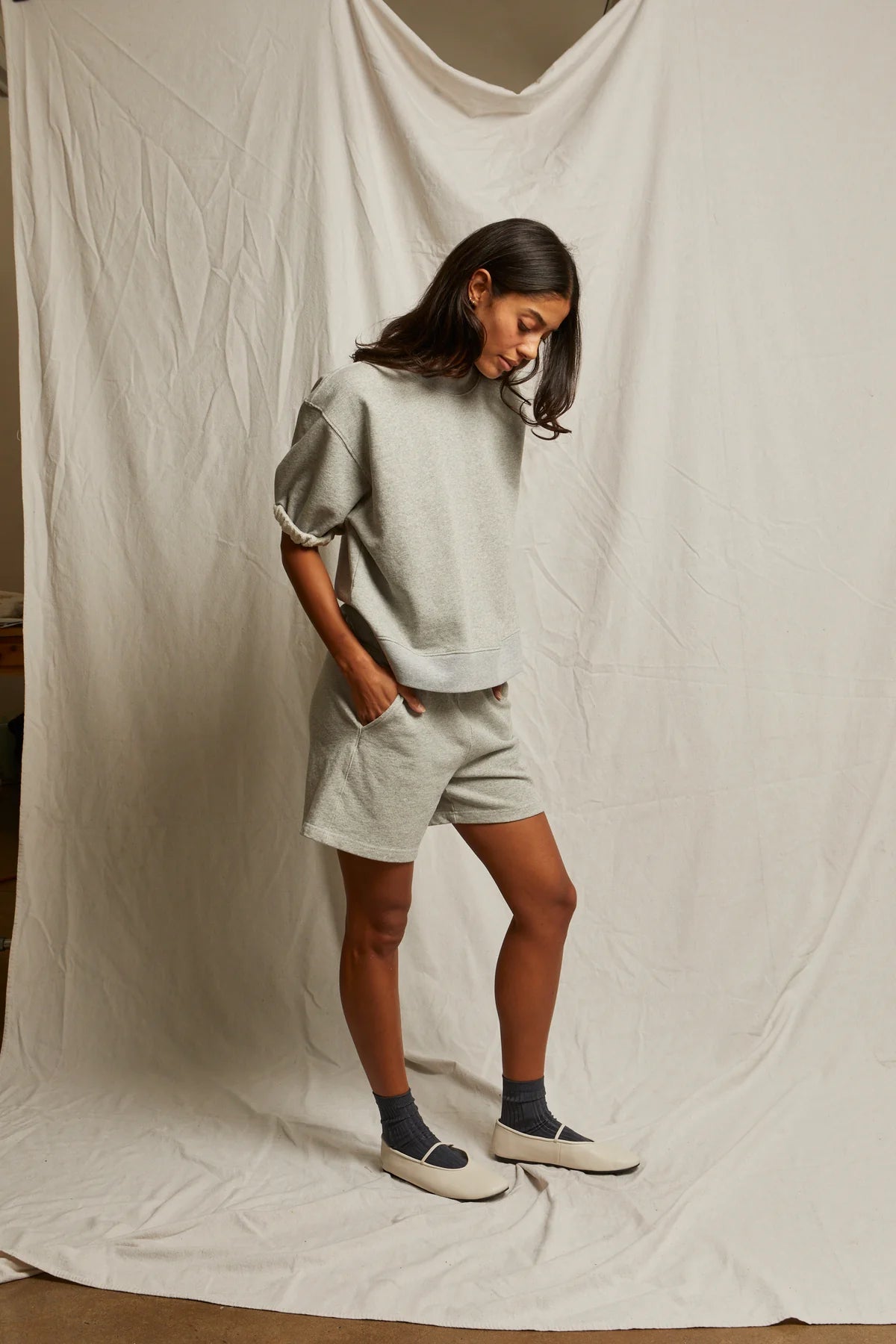 A woman stands before a cream backdrop wearing Perfectwhitetee's Rebecca SS Crew in light gray French terry cotton with matching shorts, dark socks, and white slip-on shoes. She looks down, appearing relaxed.