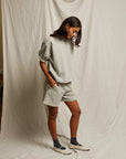 A woman stands before a cream backdrop wearing Perfectwhitetee's Rebecca SS Crew in light gray French terry cotton with matching shorts, dark socks, and white slip-on shoes. She looks down, appearing relaxed.