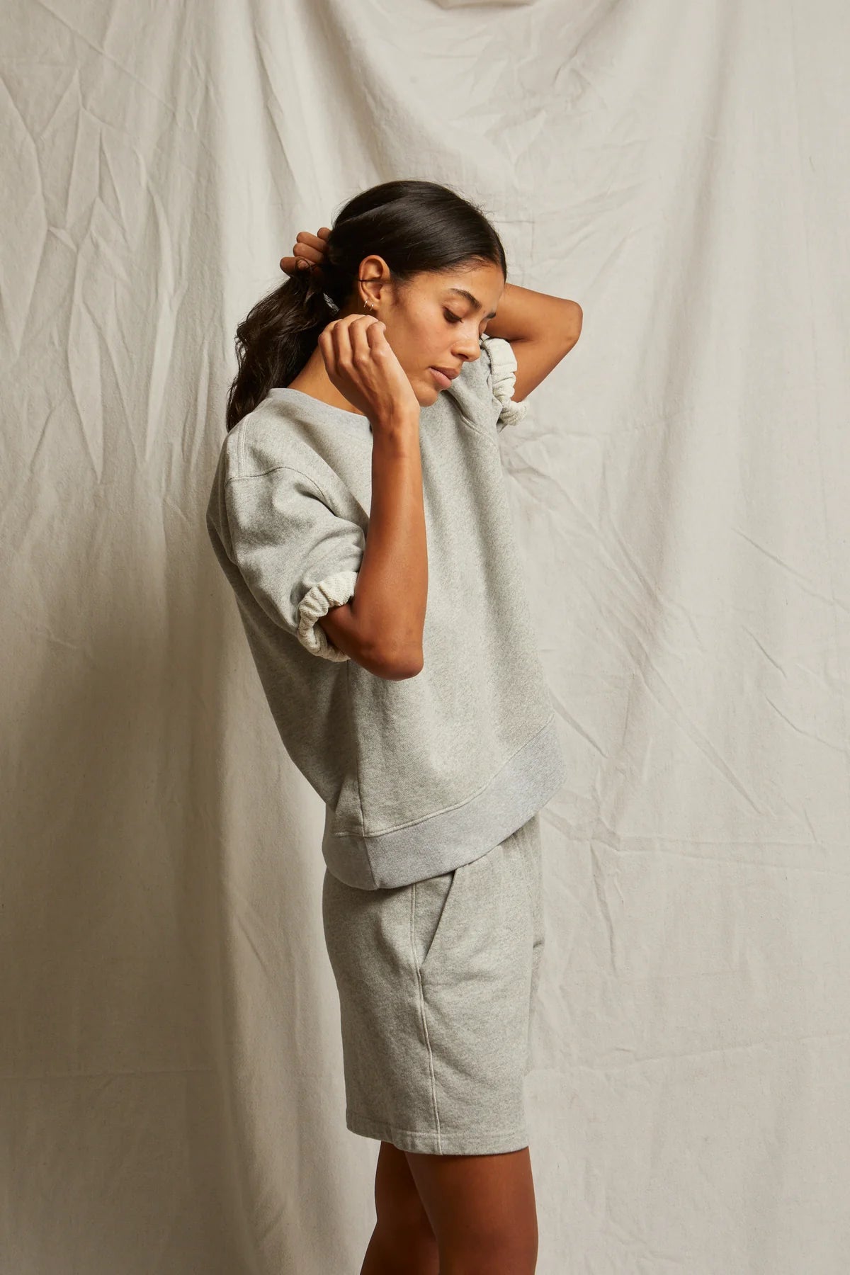 A woman adjusts her hair while wearing the Perfectwhitetee Rebecca SS Crew, a loose-fitting light gray french terry cotton sweatshirt with matching shorts, posed against a neutral wrinkled fabric backdrop.