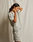 A woman adjusts her hair while wearing the Perfectwhitetee Rebecca SS Crew, a loose-fitting light gray french terry cotton sweatshirt with matching shorts, posed against a neutral wrinkled fabric backdrop.