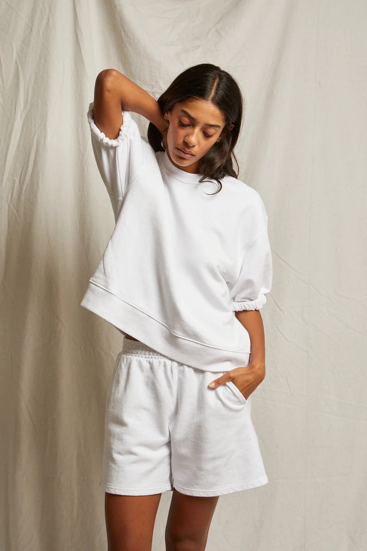 A woman poses against a light backdrop, wearing Perfectwhitetee’s Rebecca SS Crew and matching white shorts. With her left hand behind her head and right in her pocket, she gazes thoughtfully downward.