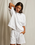 A woman poses against a light backdrop, wearing Perfectwhitetee’s Rebecca SS Crew and matching white shorts. With her left hand behind her head and right in her pocket, she gazes thoughtfully downward.