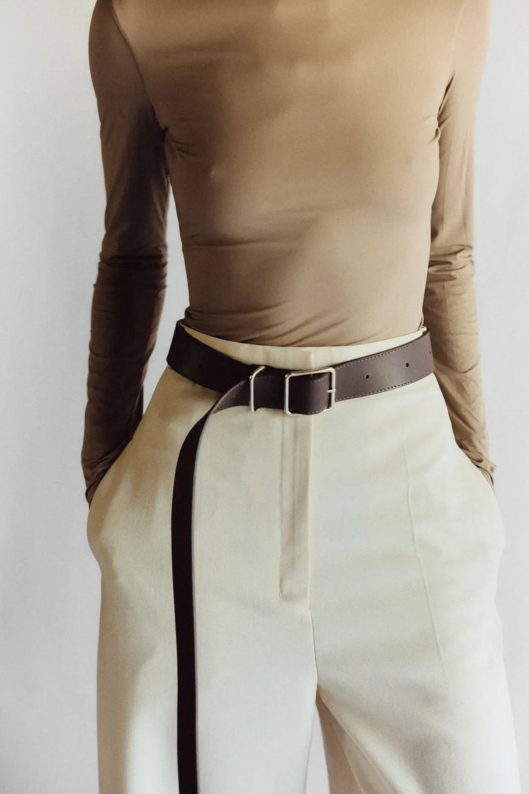 A person is pictured from the neck down, wearing a fitted beige long-sleeve top with high-waisted cream trousers. A Janessa Leone Bowe Belt in black and earth-brown regenerative leather hangs loosely. Their hands are in their pockets against a plain white background.