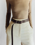 A person is pictured from the neck down, wearing a fitted beige long-sleeve top with high-waisted cream trousers. A Janessa Leone Bowe Belt in black and earth-brown regenerative leather hangs loosely. Their hands are in their pockets against a plain white background.