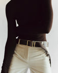 A person wears a fitted black long-sleeve turtleneck with high-waisted, light beige pants. The look is cinched by the Janessa Leone Bowe Belt, featuring custom-designed hardware and a large buckle, against a softly blurred background echoing Los Angeles elegance.