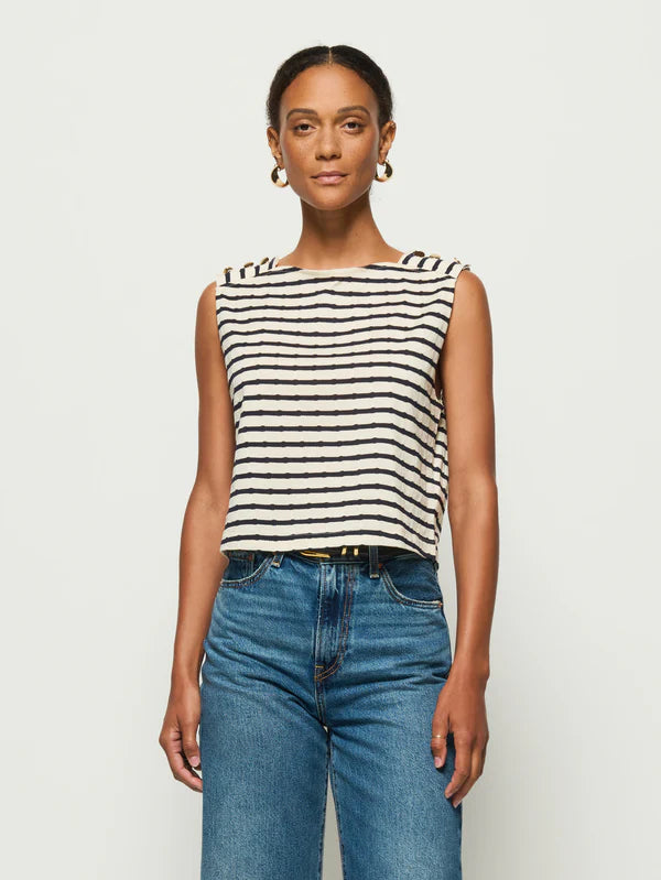 Wearing the Nation LTD Bettina Tank, with black and white stripes and gold buttons, a person pairs it with blue jeans. Their styled hair and hoop earrings frame their face while they stand against a plain backdrop, looking at the camera with a neutral expression.