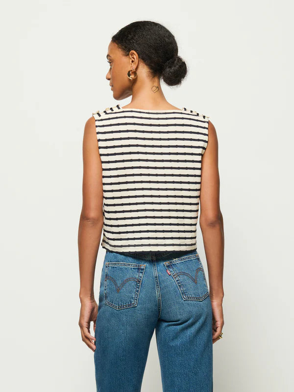 A person with short hair tied back is standing against a white background, wearing the Bettina Tank from Nation LTD, a boxy sleeveless top in black-and-white stripes made of soft Peruvian cotton, paired with blue jeans.