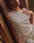 A person in a sleeveless, striped Bettina Tank by Nation LTD and matching skirt leans gracefully against a wooden wall. Adorned with hoop earrings and gold buttons, their long hair falls gently as their hand rests thoughtfully on their chest. A balcony railing is visible in the background.