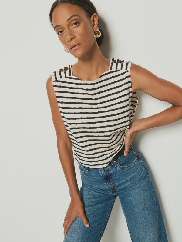 A person wears the Nation LTD Bettina Tank, a striped, sleeveless top made from soft Peruvian cotton with gold shoulder buttons, paired with blue jeans. Hoop earrings complete the look against a plain backdrop.