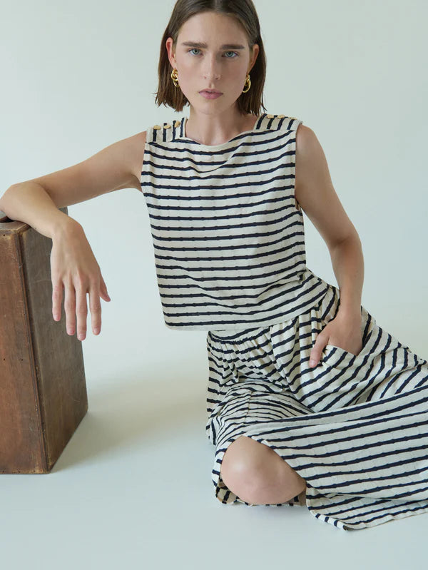 A person with short brown hair leans on a wooden box, wearing the Bettina Tank from Nation LTD—a sleeveless black and white striped dress made of Peruvian cotton. Their right hand is on the box, left hand in a pocket, against a light, neutral background.