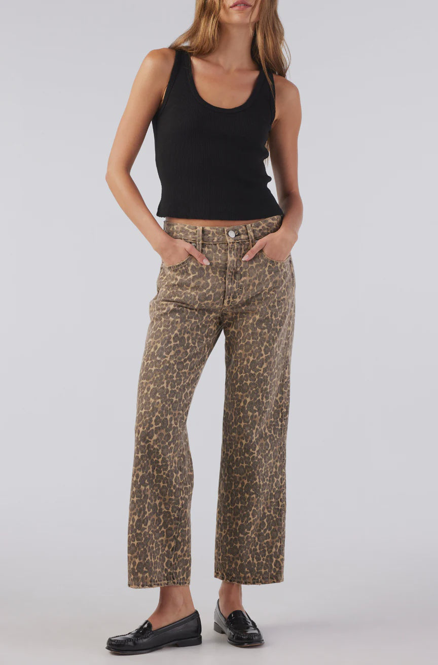 Wearing a black tank top and AMO Denim's Billie Pants in leopard print, a person stands confidently against a light background. The relaxed fit of the trousers is highlighted by their hands in the pockets, complemented by black loafers and long hair flowing freely.